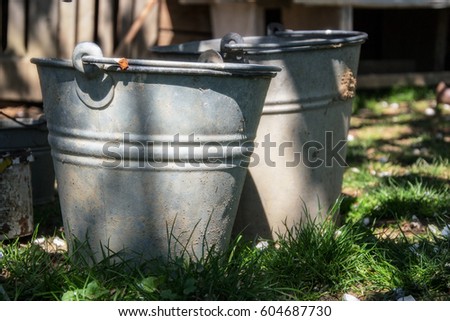 Close up of two rusty iron buckets in grass in the garden. Dirty gray metallic bucket with garbage on barnyard at sunny day. Gardening background. Containers for storing feed, trash, water, paint Royalty-Free Stock Photo #604687730