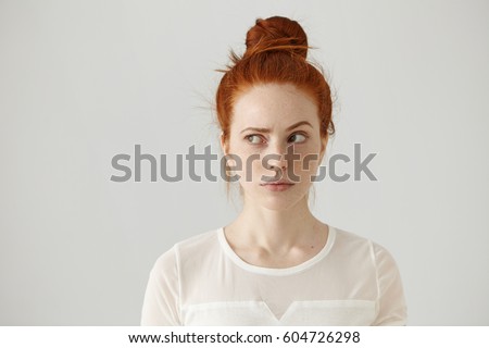 Hmm. Let me think. Studio shot of cute redhead girl with hair knot and freckles looking sideways with thoughtful and sly expression, raising one brow as if having good idea, planning something Royalty-Free Stock Photo #604726298