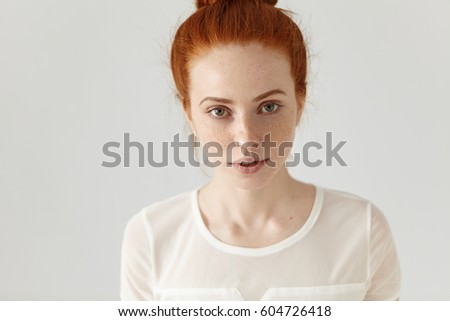 Top view of redhead pretty girl with cute subtle smile and freckles looking at camera, posing in white studio. Headshot of gorgeous young woman with clean freckled skin and ginger hair in bun