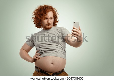 Funny overweight man with fat tummy hanging out of grey t-shirt, standing at studio wall, keeping hand on waist while posing for picture, taking selfie on mobile phone, trying to look seductive