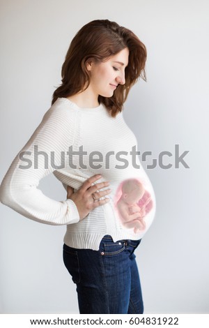 Superimposed newborn baby inside the mother's pregnant belly. a concept of development of a foetus in mother's tummy. Smiling pregnant woman posing on a white wall background. Pregancy concept. Royalty-Free Stock Photo #604831922