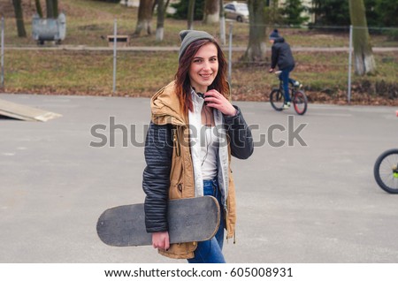 Portrait of of a young happy girl holding a skateboard. Skate park cyclist in the background
