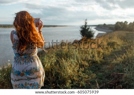 Back view of a young woman wanderer is making photo with portable tablet camera during her vacations in village meadows with great river