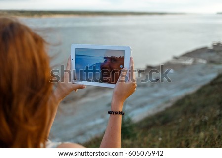 Back view of a young woman wanderer is making photo with portable tablet camera during her vacations in village meadows with great river. In reflection of the screen the smiling face of the girl is