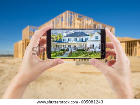 Female Hands Holding Smart Phone Displaying Photo of House with Unfinished Home Construction Behind.