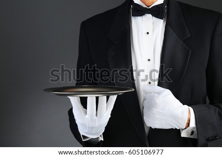 Closeup of a tuxedo wearing waiter holding a silver tray in front of his body. Horizontal format on a light to dark gray background. Royalty-Free Stock Photo #605106977