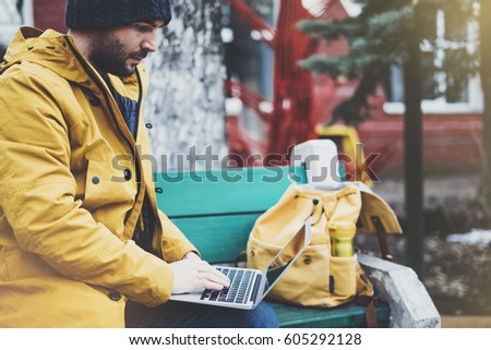 Hipster with yellow backpack, jacket, cap, thermos cup of coffee using computer open laptop in spring street outdoor, tourist man typing on keyboard, traveler connect internet, freelance