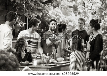 Young people having fun on the terrace, drinking beers and chatting while their friends serve the meat roasted on the bbq. Black and white picture