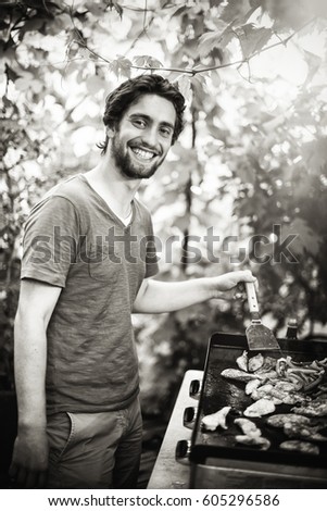 Looking at camera. A young man grilling meat on a bbq for his friends. Black and white picture