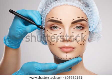 Pretty girl with dark eyebrows wearing blue medical hat at studio background, doctor's hands wearing blue gloves drawing perforation lines on face, plastic surgery concept. Royalty-Free Stock Photo #605382959