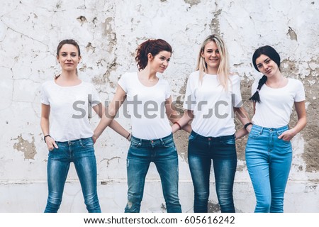 Group of four young diverse girls wearing blank white tshirt and jeans posing against rough street wall, fashion urban clothing style, mockup for t-shirt print store Royalty-Free Stock Photo #605616242