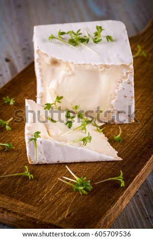 Vertical photo with unusual camembert. Cheese with cube shape with spilled arugula on vintage chopping board with cut off portion and on worn color wooden table with various colors. 