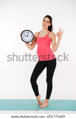 Full length portrait of a happy young pregnant woman holding an alarm clock and showing okay gesture isolated on white background