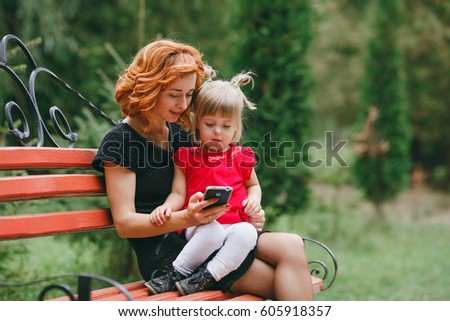 mother and her little daughter sitting on a park bench and enjoy the phone