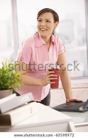 Office worker leaning at desk holding coffee to go cup, looking up, smiling.?