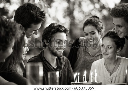 A group of young people gathered on a terrace in a garden to celebrate the birthday of one of their friends. A young man is blowing candles on the cake. Black and white picture