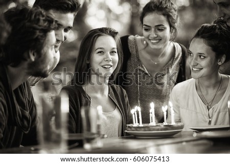 A group of young people gathered on a terrace in a garden to celebrate the birthday of one of their friends. A young woman is blowing candles on the cake. Black and white picture