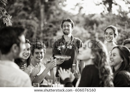 A group of young people gathered on a terrace to celebrate the birthday of one of their friends. A young man holding a cake with candles on it. Black and white picture.