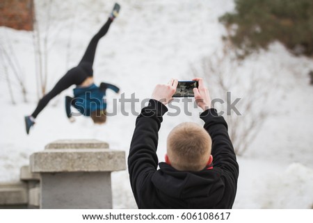Teenager makes photo on smartphone of acrobatic jump girl in winter city park - parkour concept