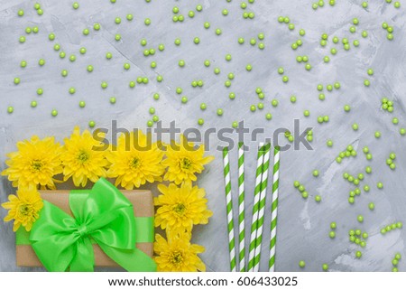Gift box craft paper with green bow ribbon, yellow chrysanthemum flowers, 
Stripped green paper straws for cocktails, decorating sugar balls on grey 
concrete background. Place for copyspace.