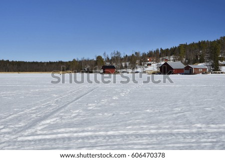 Snow and ice and snowmobile tracks and a little fishing village with red boathouses against a blue sky, picture from the North of Sweden.