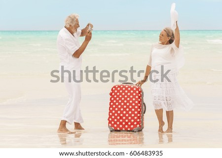 Happy mature Caucasian retired couple in white posing on vacation beach with camera and travel luggage