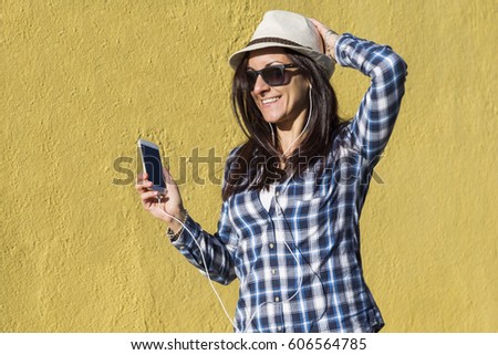 happy beautiful young woman listening to music and having fun over yellow background. She is wearing hat and modern sunglasses. lifestyle