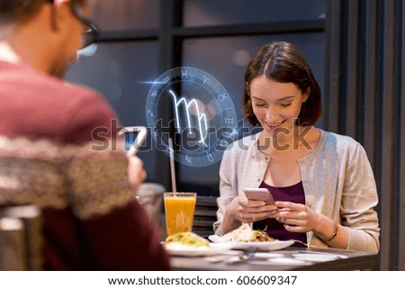 technology, astrology, horoscope and people concept - happy couple or friends with smartphones and scorpio zodiac sign having dinner at restaurant 