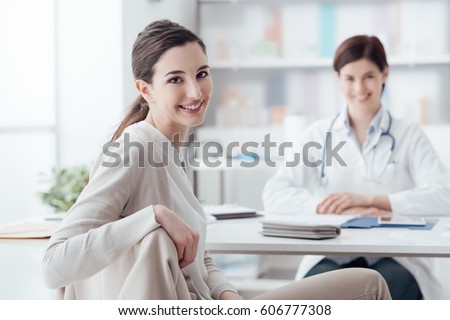 Smiling patient receiving a medical consultation and looking at camera, the female doctor is sitting at desk on the background