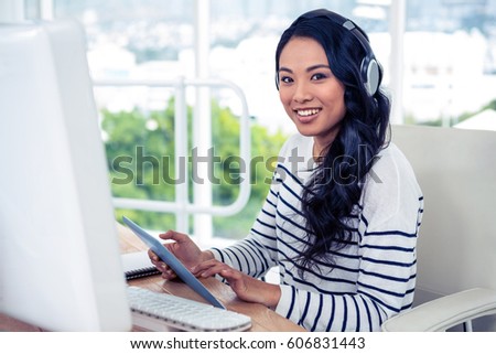 Smiling Asian woman with headphones using tablet and looking at the camera in office