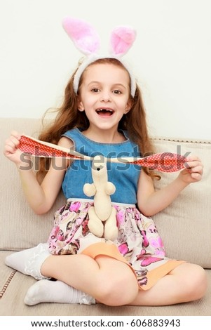 Cute little girl with bunny ears and cuddly toy sitting on sofa at home.