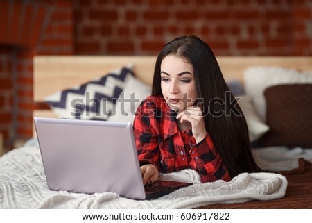 Pretty woman relaxing on the bed in her room and chatting on her laptop. Indoors. Online friendship and social network concept.
