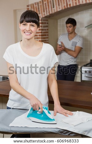 Woman ironing a shirt while man using mobile phone in background