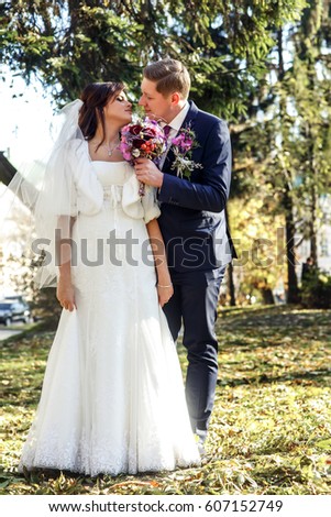 Groom with original violet wedding bouquet stands behind bride in autumn park
