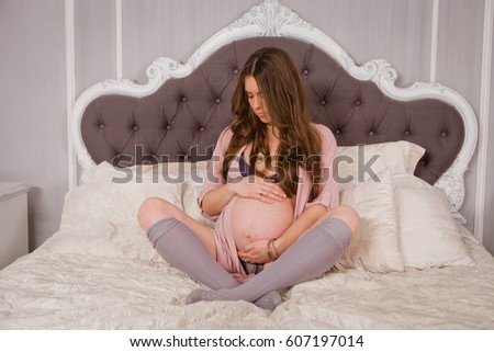 A beautiful young pregnant expectant mother sits in her underwear and pinoyar in a lotus pose and arms her belly on a double bed in a white bedroom. Beautiful makeup and styling on long blond hair