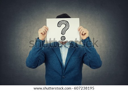 Businessman covering his face using a white paper with drawn question mark, like a mask, for hiding his identity. Isolated gray wall background. Royalty-Free Stock Photo #607238195