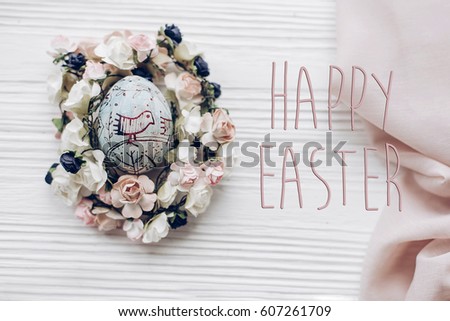 happy easter text greeting card sign on  easter egg with chick ornaments in floral nest with flowers on rustic white wooden desk background. space for text, top view. soft light