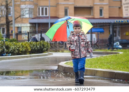 Little boy playing in rainy summer park. Child with colorful rainbow umbrella, waterproof coat and boots jumping in puddle and mud in the rain. Kid walking in autumn shower Outdoor fun by any weather.