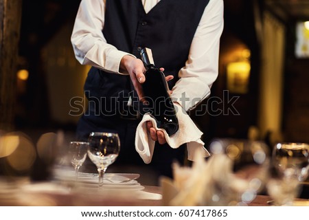 The waiter in uniform with a white towel offers visitors wine in restaurant Royalty-Free Stock Photo #607417865