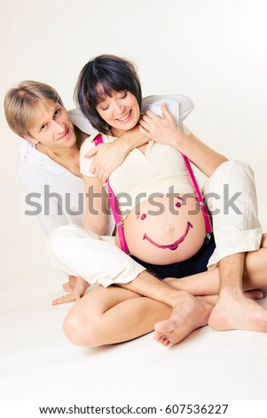 Husband hugging his pregnant wife sitting on the floor. Woman's belly is painted with a smile