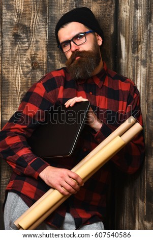 Bearded man, long beard. Brutal caucasian serious unshaven hipster holding laptop and craft paper rolls in red black checkered shirt with hat and glasses on brown vintage wooden studio background