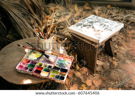 Oil paints and paint brushes in plastic can put near palette in the garden,practices drawing picture. 