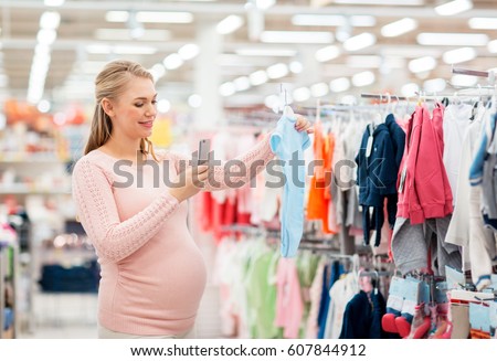 pregnancy, people and shopping concept - happy pregnant woman taking picture of blue baby bodysuit with smartphone at clothing store