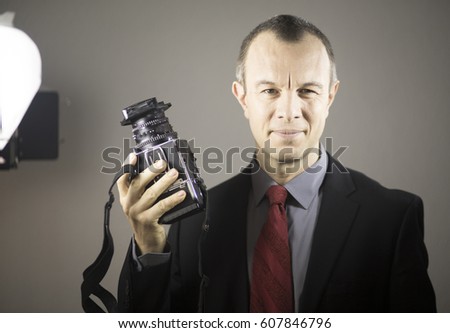 Photographer in studio in suit with camera aged in 40's against plain studio portrait background.