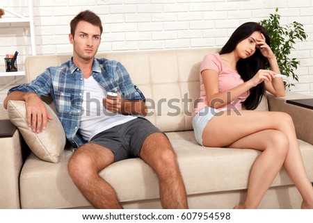 Upset woman is looking in pregnancy test. Indifferent man watching TV.