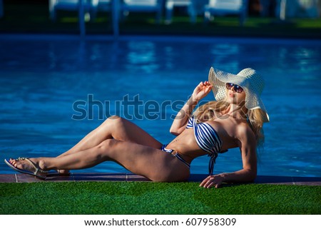 A beautiful young girl is lying on the ledge of the pool. She is wearing a pretty swimsuit, sunglasses and hat. She is sunbathing.