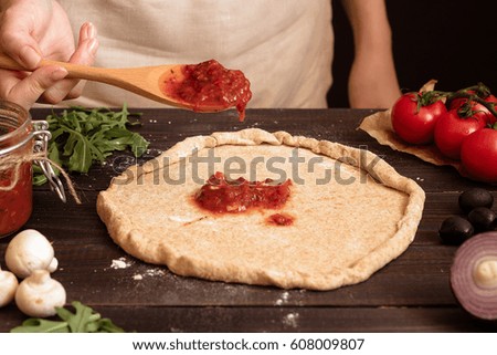 Cooking pizza. Hands adding fresh tomato sauce to pizza dough. Pizza ingredients on the wooden table.