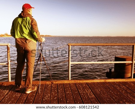 Tall nature photographer at tripod taking picture on the wooden pier in wharf at sunset time. 