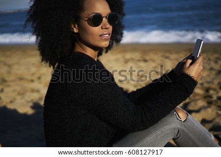 Afro american hipster girl with curly hair and in eyewear taking photo using smartphone recreating sitting on beach enjoying weekends, charming female making selfie for updating profile picture