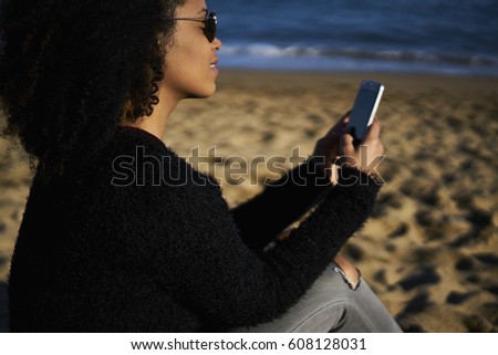 Charming attractive afro american female traveler in glasses making selfie using smartphone to share with followers in social networks sitting on sea coast enjoying sunny weather and weekends